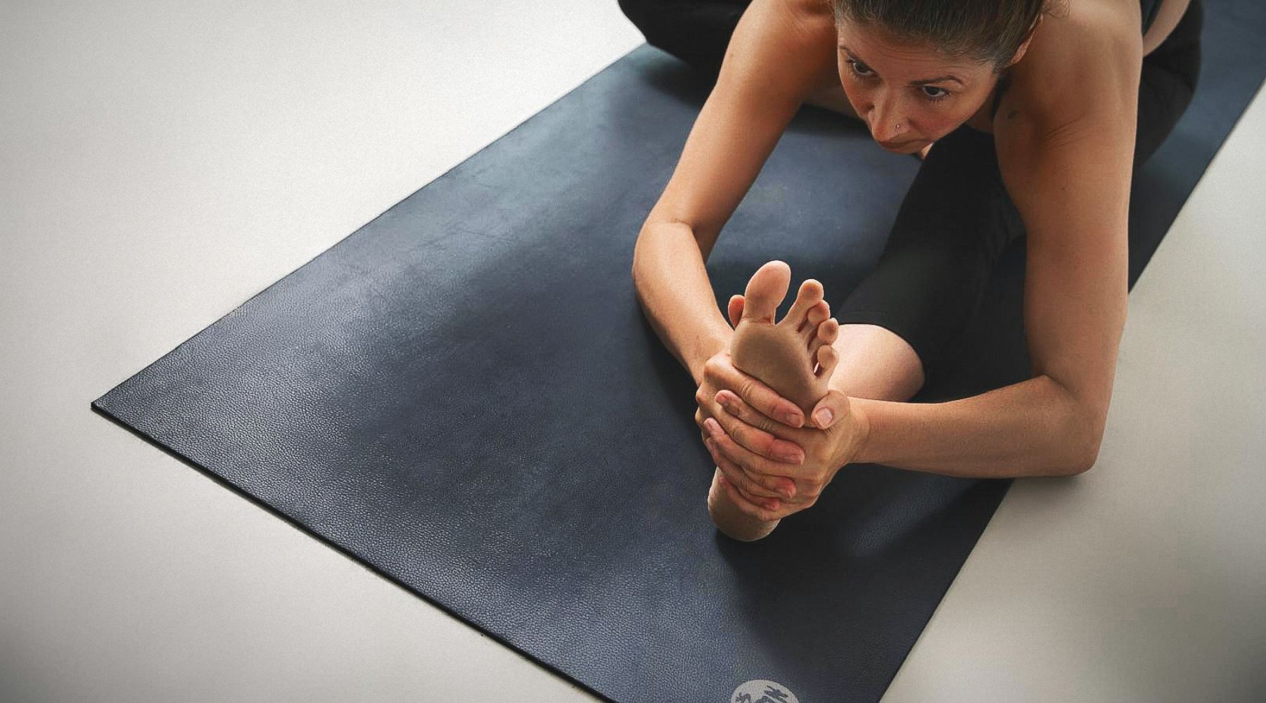 A person stretches on a black yoga mat, grasping their foot with both hands, focusing on flexibility and balance.