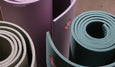 Three rolled yoga mats in muted colors: lavender, olive green, and deep teal, showcasing their textured surfaces and circular forms.