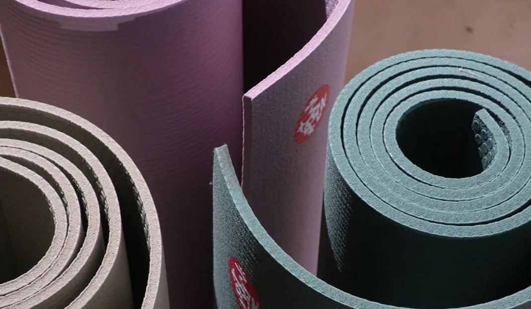 Three rolled yoga mats in muted colors: lavender, olive green, and deep teal, showcasing their textured surfaces and circular forms.