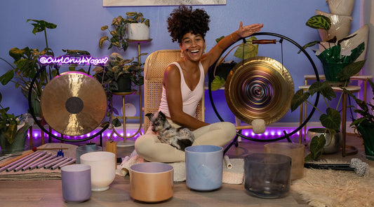 A person with curly hair sits cross-legged among various sound healing instruments, plants, and a small dog, in a serene atmosphere.