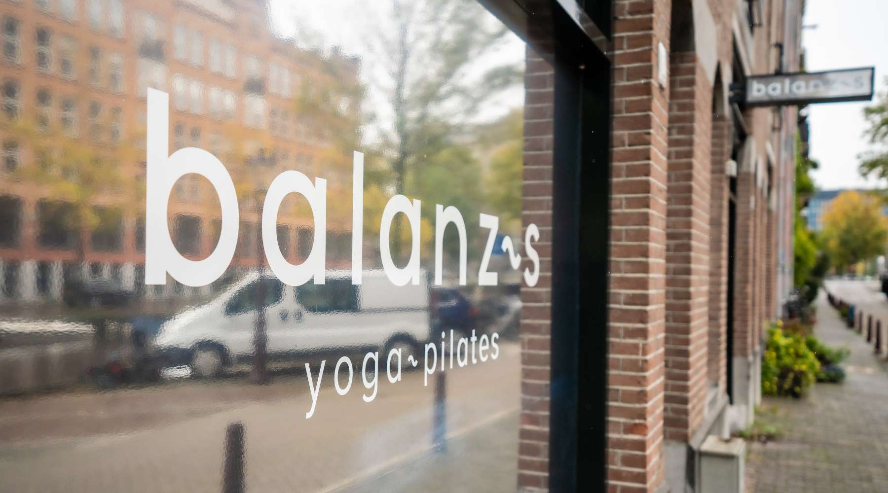 A close-up of a storefront window displaying the word "balanzs" and the phrases "yoga" and "pilates," with a blurred city view behind it.