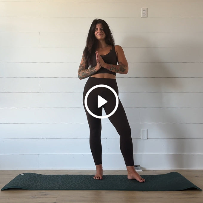 Tattooed woman in black workout clothes stands barefoot on a Pilates mat with hands together; play button overlay indicates a video.