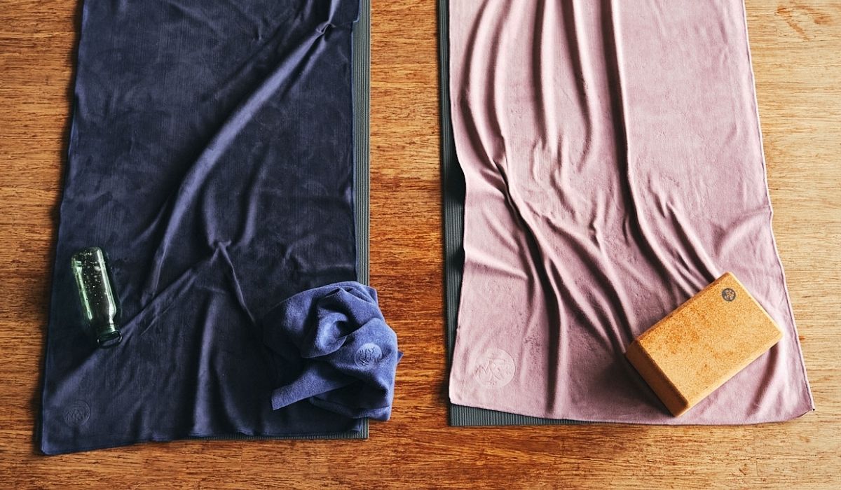 Two hot yoga towels, one navy with a water bottle, the other purple with a cork block, placed on a wooden surface.