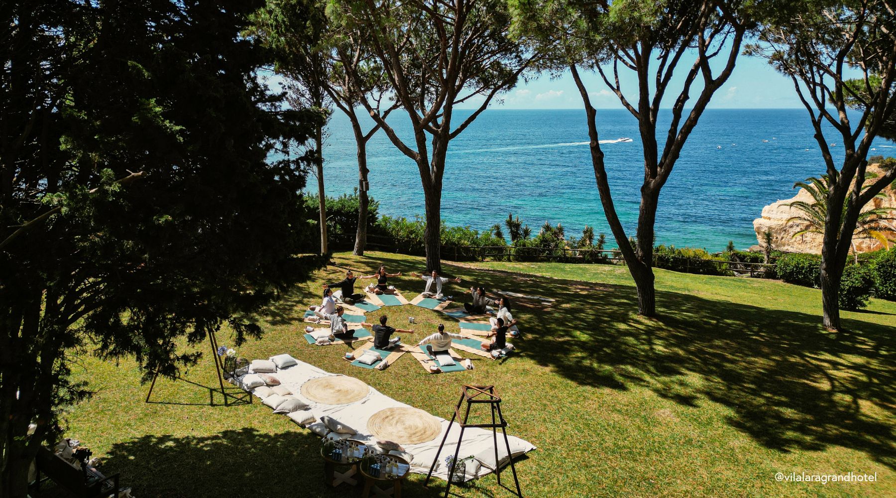 A group of people meditating on manduka yoga mats in a circular formation under trees, overlooking a serene blue ocean.