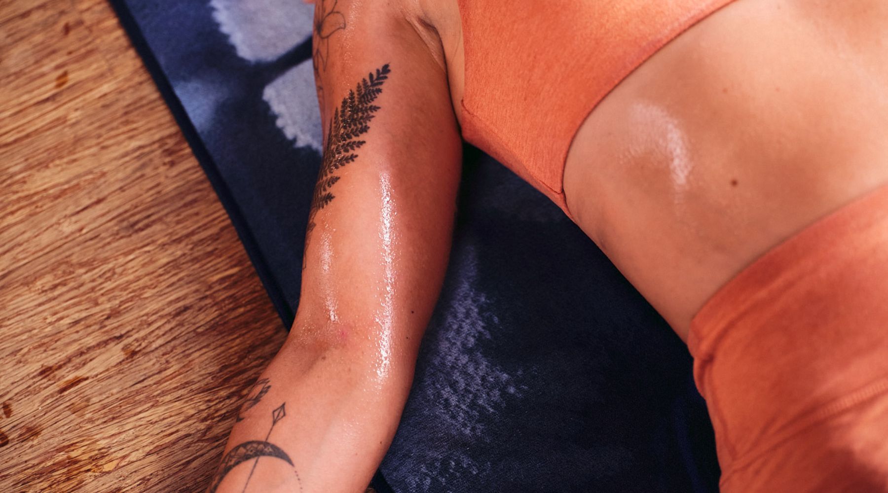 A close-up of a woman's arm with tattoos, glistening with sweat, resting on a blue yoga towel. She's wearing an orange workout top.