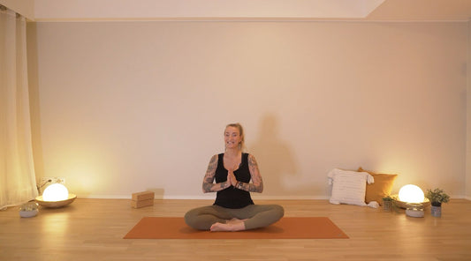A yoga teacher sits cross-legged on a yoga mat in a serene room, hands in prayer position, surrounded by soft lighting and decor.