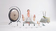 A person sits cross-legged on a yoga mat, holding instruments in each hand, surrounded by singing bowls, a gong, and decorative cushions.