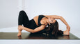 A woman in athletic wear performs a challenging yoga pose on a black Manduka yoga mat, showcasing flexibility and balance against a light background.