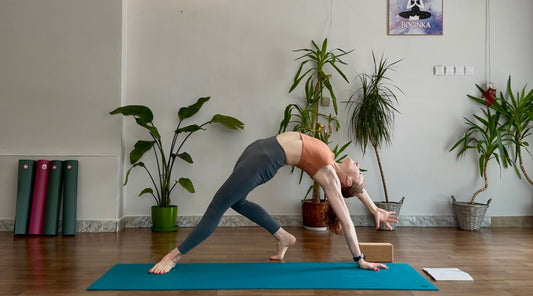 A woman in yoga clothing performs a yoga pose on a manduka yoga mat surrounded by indoor plants, capturing a moment of balance and flexibility.
