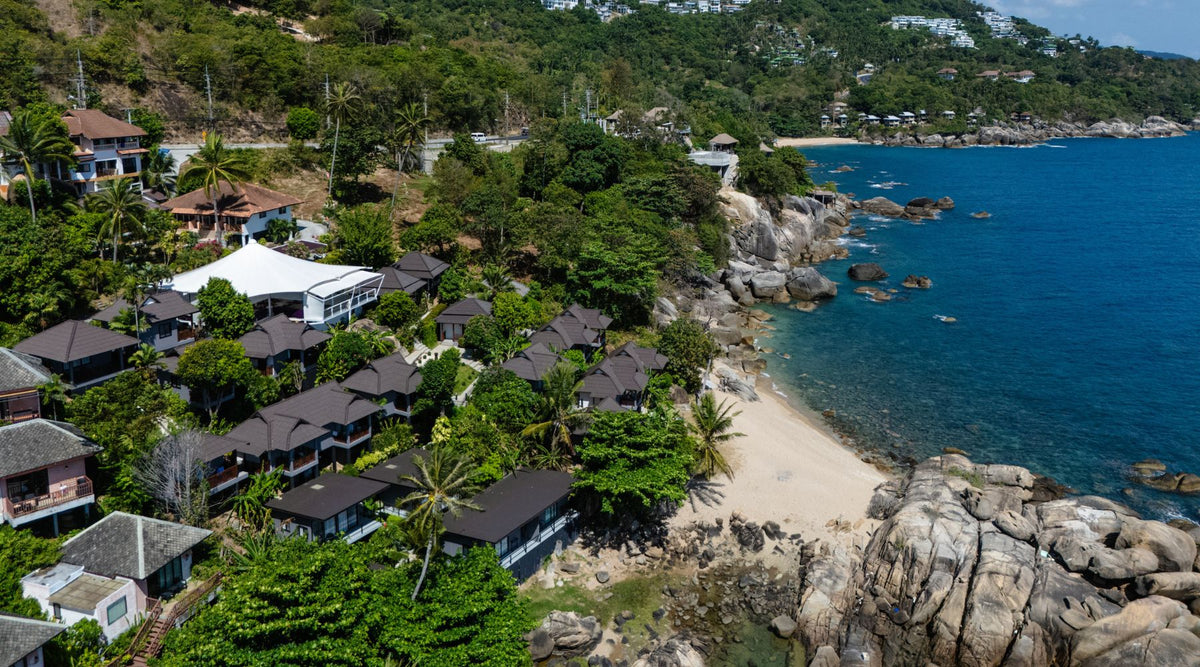 Aerial view of Vikasa a coastal center with lush greenery, beach, rocky shoreline, and villas nestled among palm trees by the blue sea.