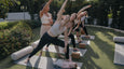 Raisa guides four pregnant and postpartum students with their babies into extended side angle pose in an outdoor yoga class.