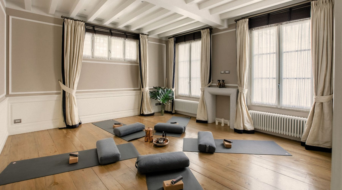 A serene yoga studio with gray mats, bolsters, and blocks arranged on wooden floors, framed by natural light and elegant curtains.