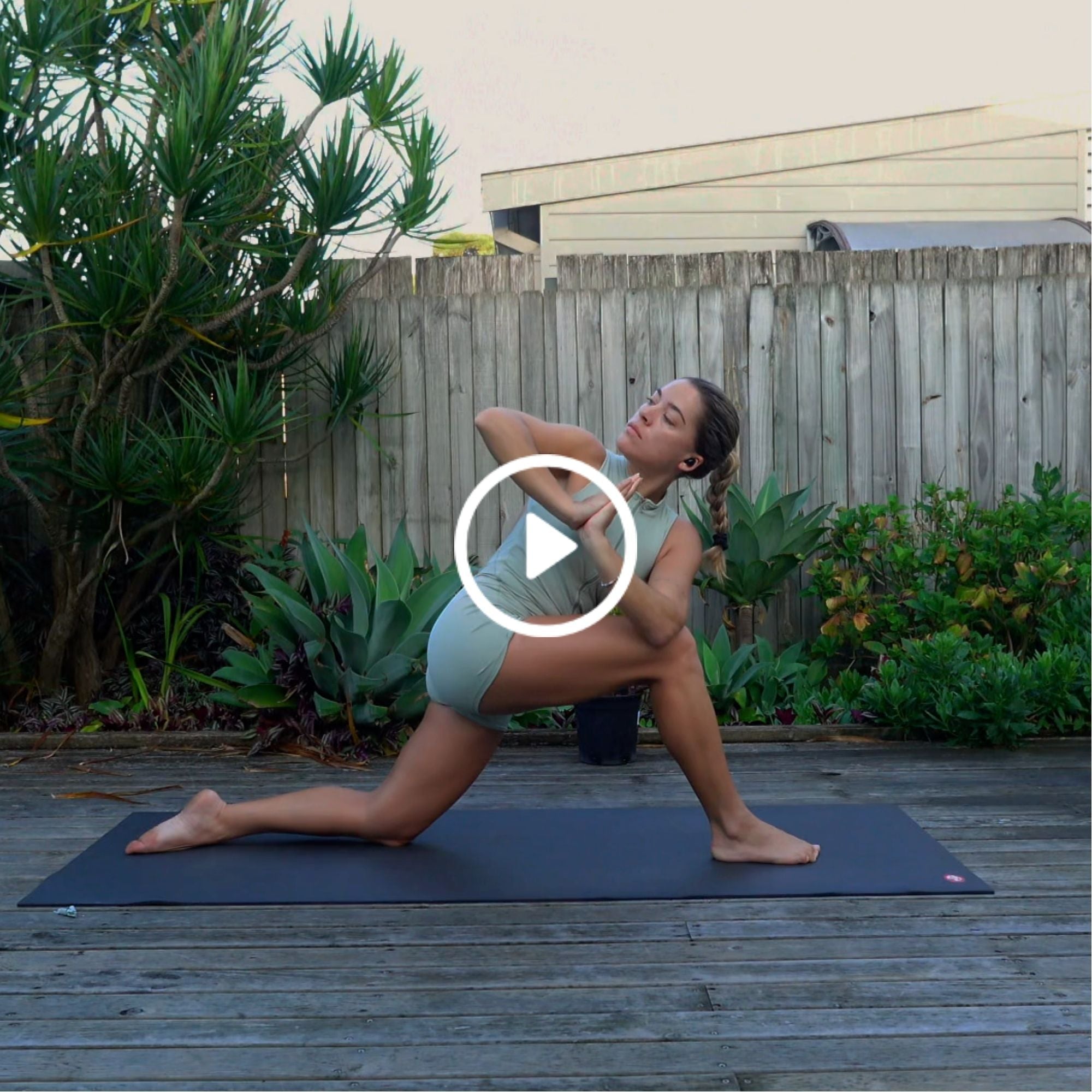 Woman in activewear practices yoga twist pose on mat outdoors on wooden deck with plants and fence in background.