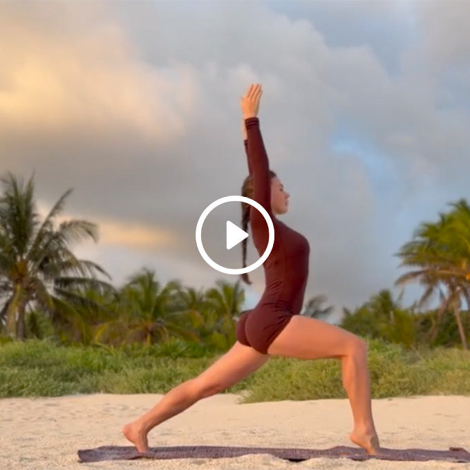 Woman in a maroon outfit practices yoga in a high lunge pose on a beach with palm trees and cloudy sky in the background.