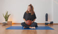 A person sits cross-legged on a blue yoga mat, holding a basketball, surrounded by plants and yoga equipment in a serene space.