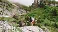A woman performs a challenging yoga pose on a large rock, surrounded by lush green mountains and misty weather.