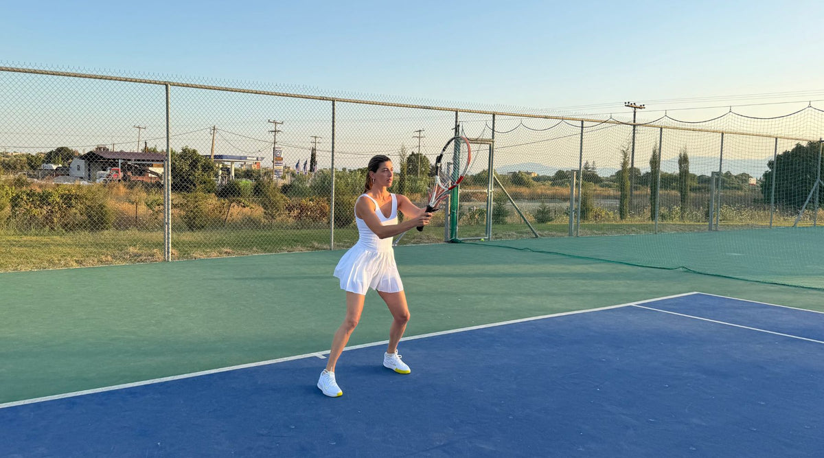 A tennis player in a white dress serves on a court, with a green landscape and blue sky in the background during sunset.