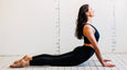 A woman performs the cobra yoga pose on a wooden floor, wearing a black sports bra and leggings, with a light-colored wall behind her.