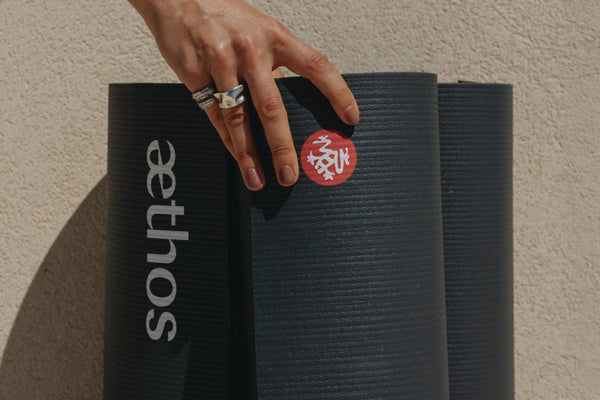A close-up of a hand resting on a rolled Mnduka black yoga mat with the brand logo. Another rolled mat with a custom brand name 'aethos' and a textured beige wall are in the background.