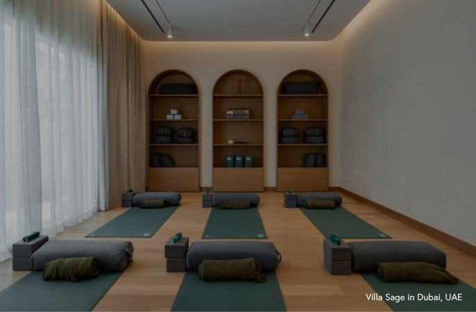 A yoga studio room with multiple dark green Manduka yoga mats arranged on a light wood floor, each with a dark gray Manduka bolster and rolled mat underneath. Built-in wooden shelves with folded Manduka blue mats are in the background, with sheer white curtains on the left.