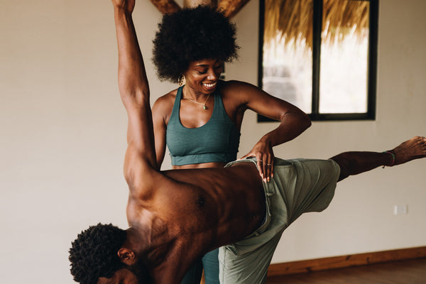 A fitness lifestyle scene showing a man in sage green shorts and a woman in a teal sports bra performing a yoga pose together in a brightly lit room.