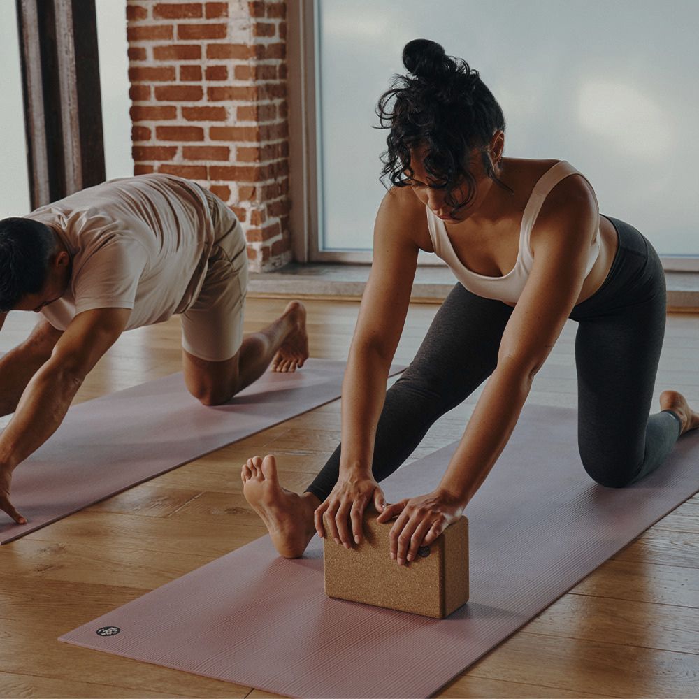 Three people practicing yoga in a bright studio with wooden floors and brick columns, using Manduka pink mats and a Manduka cork yoga block.